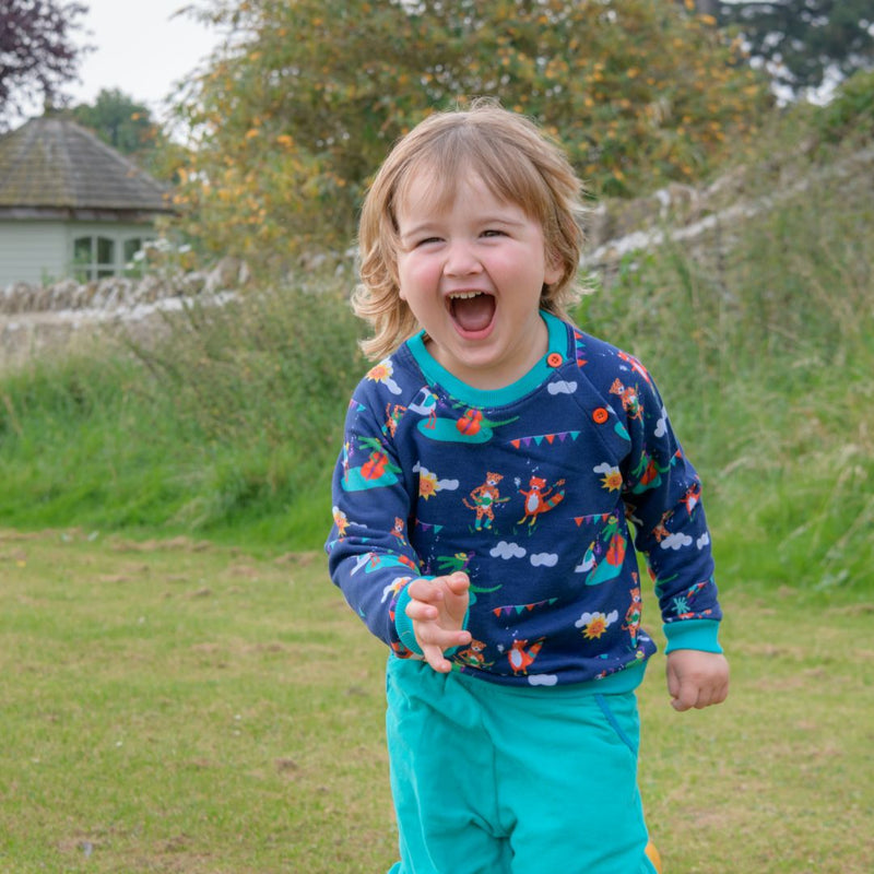 Smiling boy running outside towards the camera, wearing Ducky Zebra sweatshirt and cords. The jumper is blue with a fun print of a cheetah, fox, duck and crocodile playing music together. The jumper has a festival vibe. It has two orange buttons on the shoulder for easy dressing and contrasting turquoise cuffs.