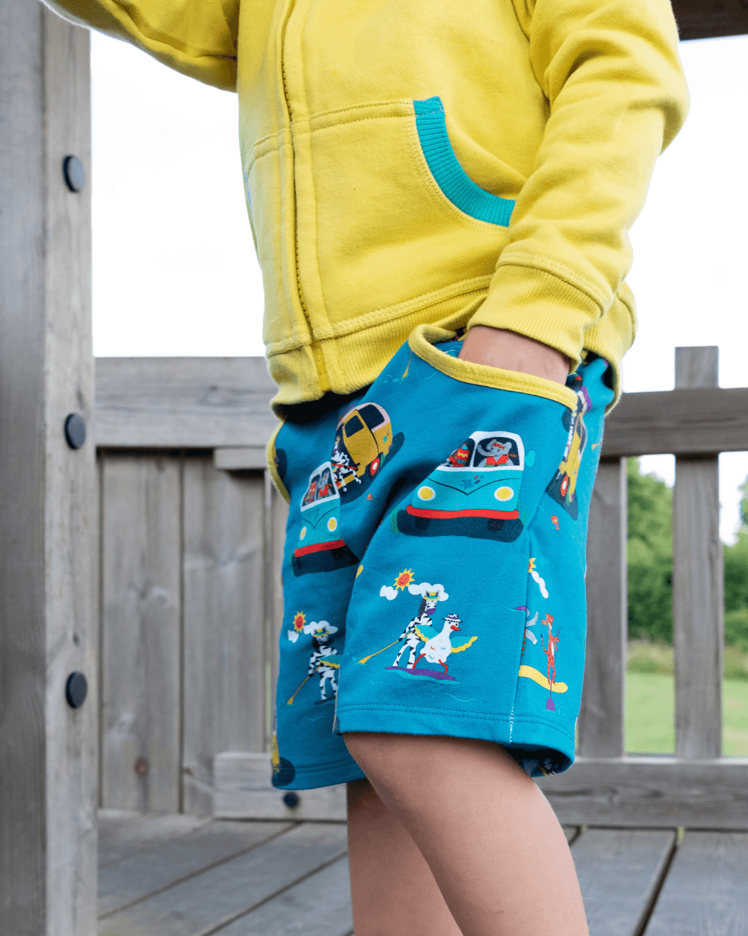 Child wearing a yellow hoodie and teal shorts with a campervan and paddleboard print, yellow waistband and two big pockets