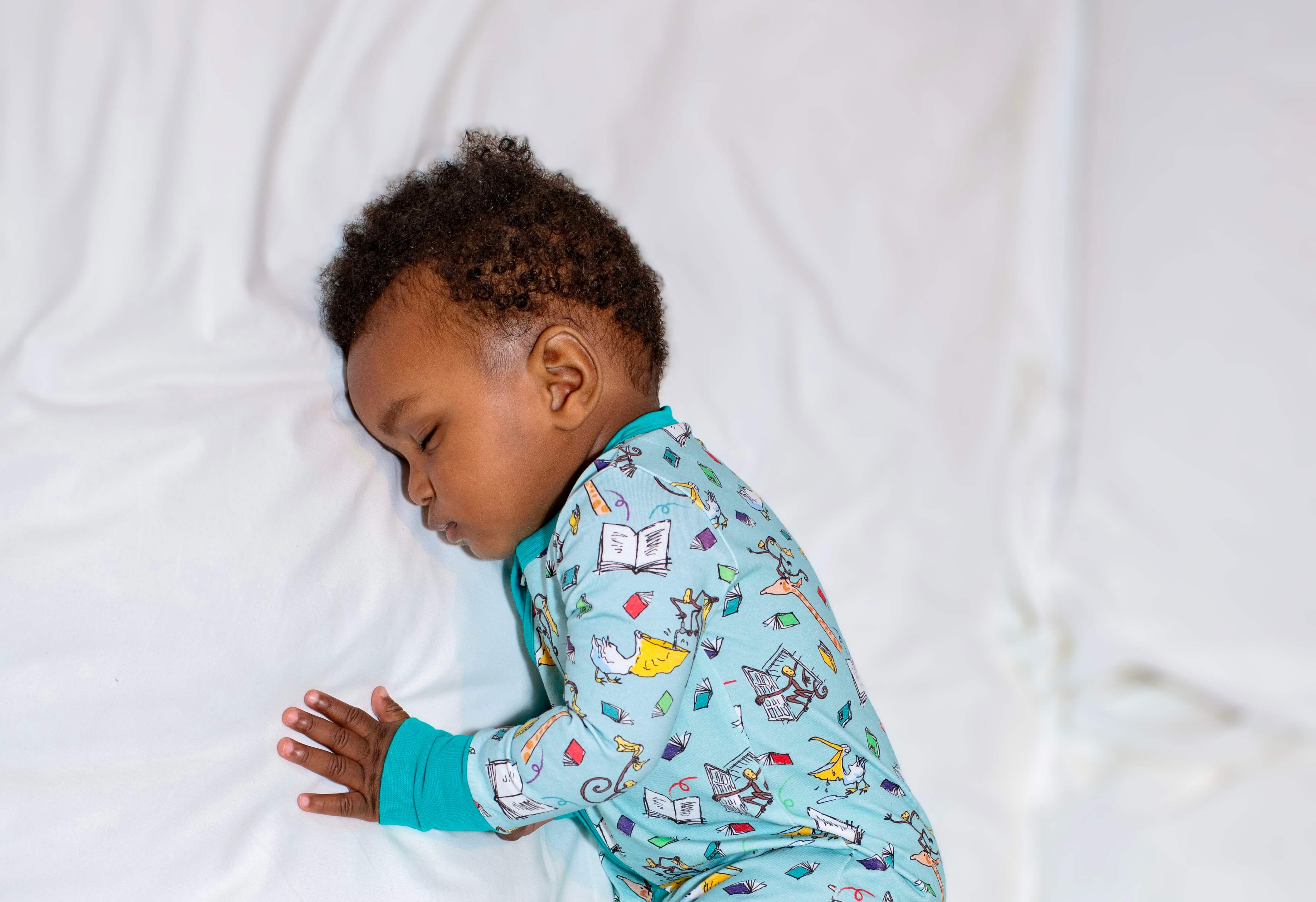 Child wearing colorful pajamas with a pattern of animals and books, sleeping on a white blanket.
