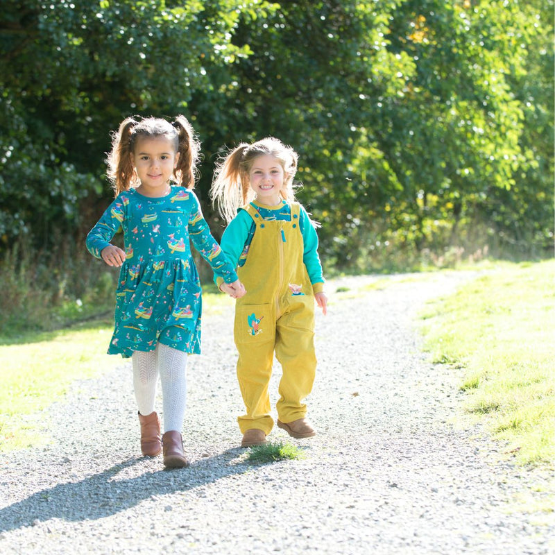 Two children walking hand in hand along a sunny pathway. They're both wearing Ducky Zebra clothes - one in a teal skater dress with a fun highland cow repeat print. The other is wearing unisex yellow cord dungarees.