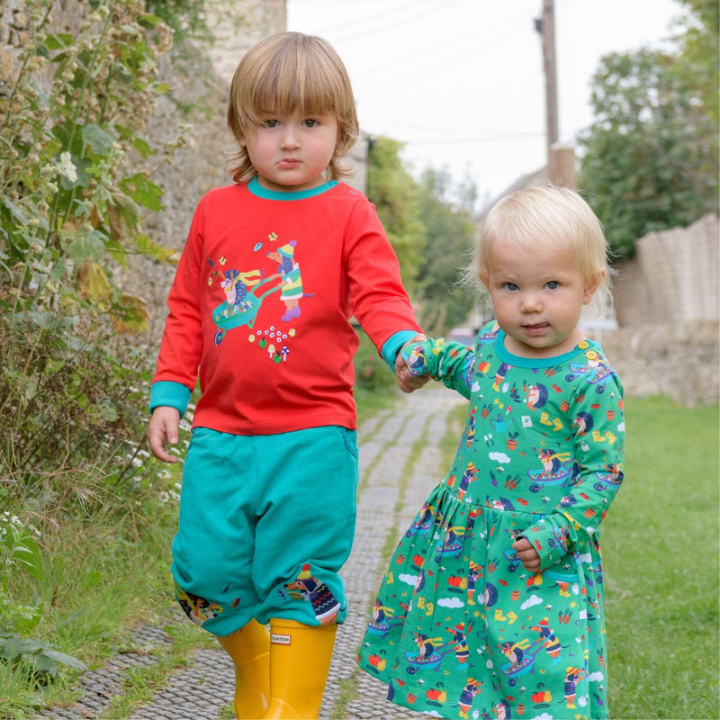 Two children holding hands, walking outside. One child is wearing a Ducky Zebra red t-shirt with a print of a dog and hedgehog. He's wearing coordinating turquoise trousers and yellow wellies. The other child is wearing a long sleeve Ducky Zebra skater dress with a repeat print of a dog and hedgehog playing together. 