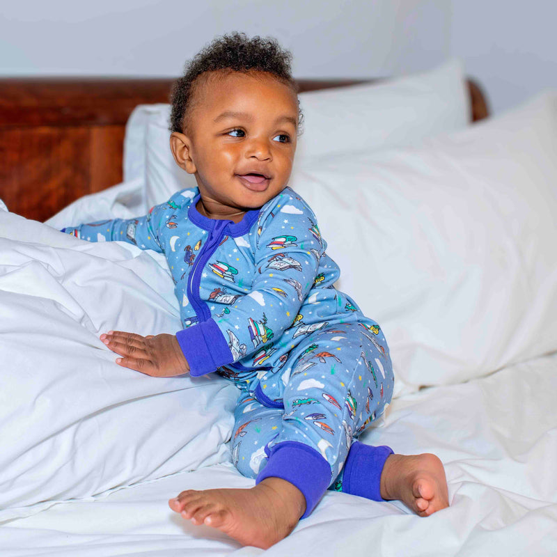 Smiling baby lying in bed wearing a zip-up sleepsuit with a fun print featuring animals from Roald Dahl stories, including the newt from "Matilda," Alfie the tortoise from "Esio Trot," and Muggle-Wump the monkey from "The Twits". 