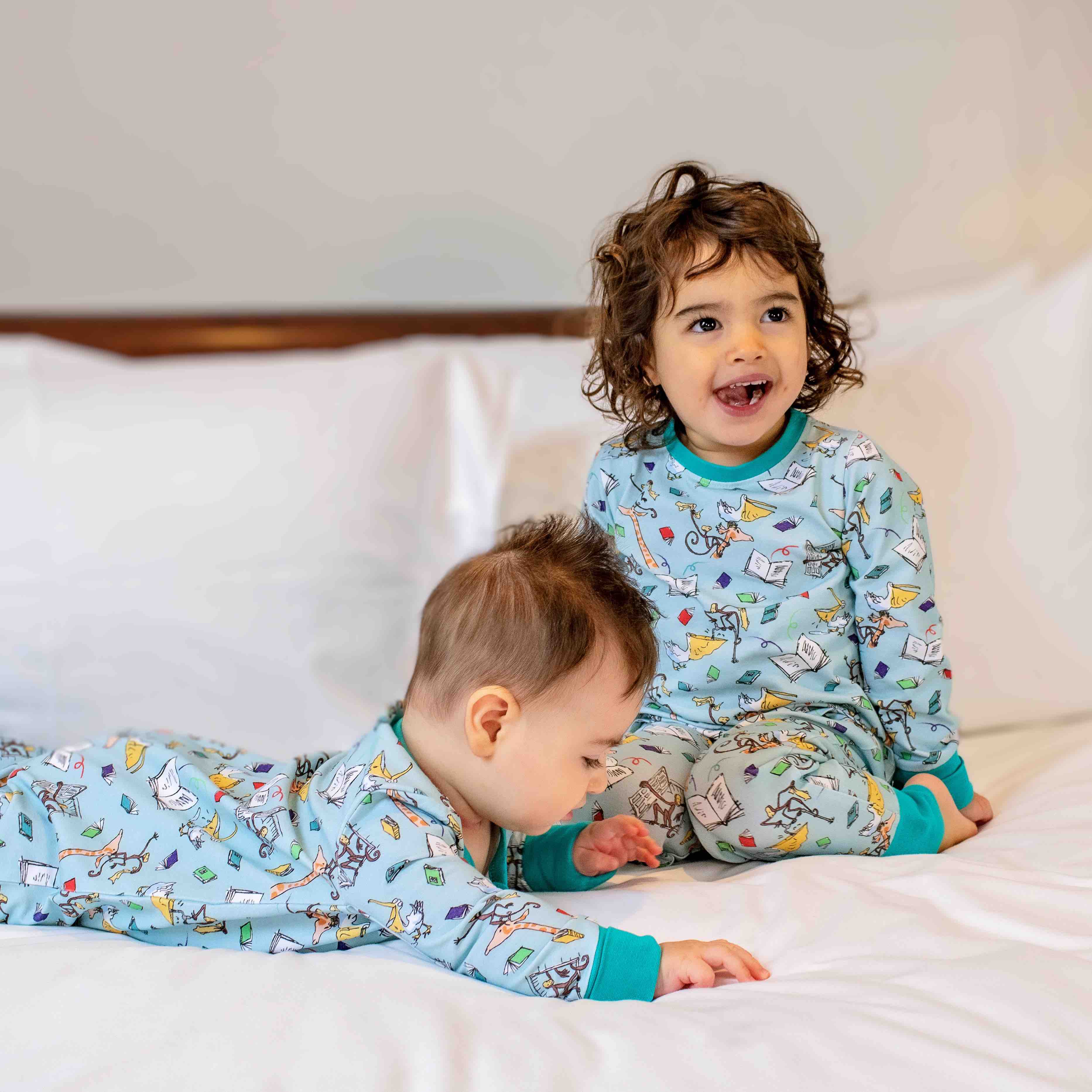Toddler and baby sitting together on a bed, with the toddler wearing Ducky Zebra Roald Dahl PJs and the baby wearing a twinning organic cotton sleepsuit. 