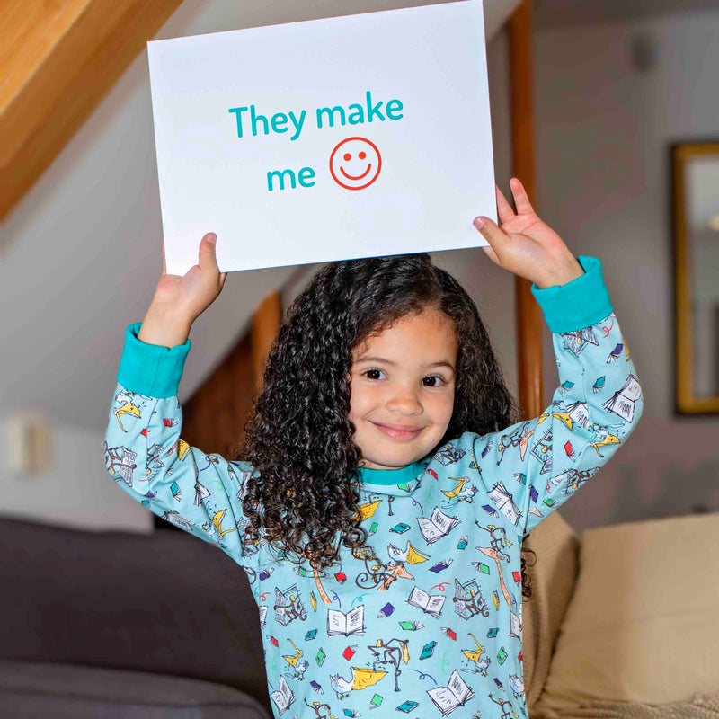 Smiling girl wearing Ducky Zebra x Roald Dahl organic cotton PJs, which feature images from The Giraffe and the Pelly and Me. The girl is holding a sign saying 'They make me happy'