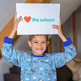 Boy wearing Ducky Zebra x Roald Dahl Pyjamas holding a sign saying "I love the colours". The pyjamas include Quentin Blake's illustrations reading stories, with a vibrant purple trim