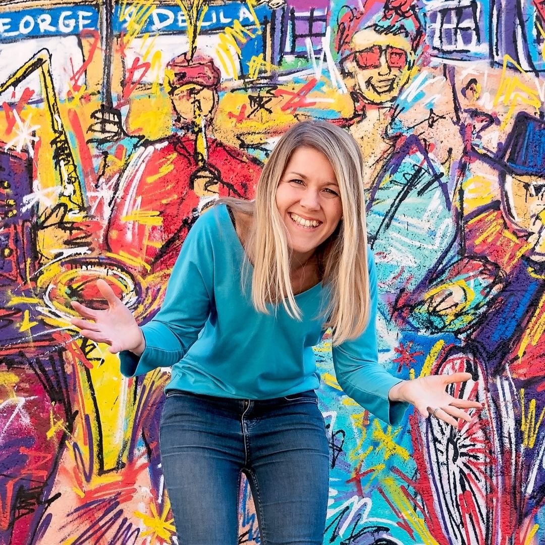 Image of Sally, founder of Ducky Zebra, smiling, with her hands out and open, in front of a brightly painted wall 