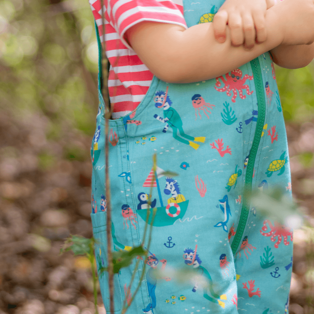 A close up of a child wearing Ducky Zebra short dungarees with a light blue background and an 'under the sea' themed print