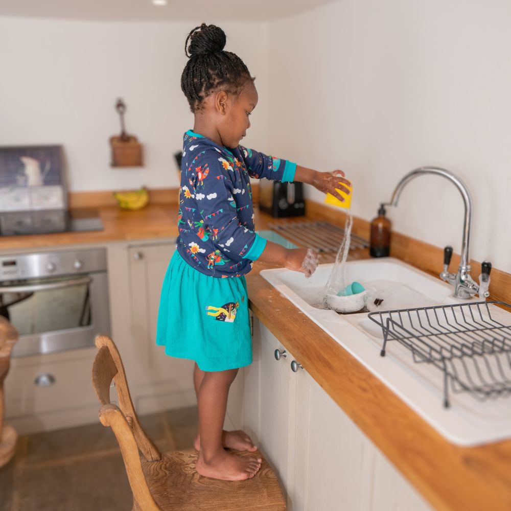 Image of a girl playing with water wearing a Ducky Zebra turquoise cord sirt with a hedgehog appliqué peaking out of the pocket. The girl is also wearing a Ducky Zebra sweatshirt with a fun festival repeat print.