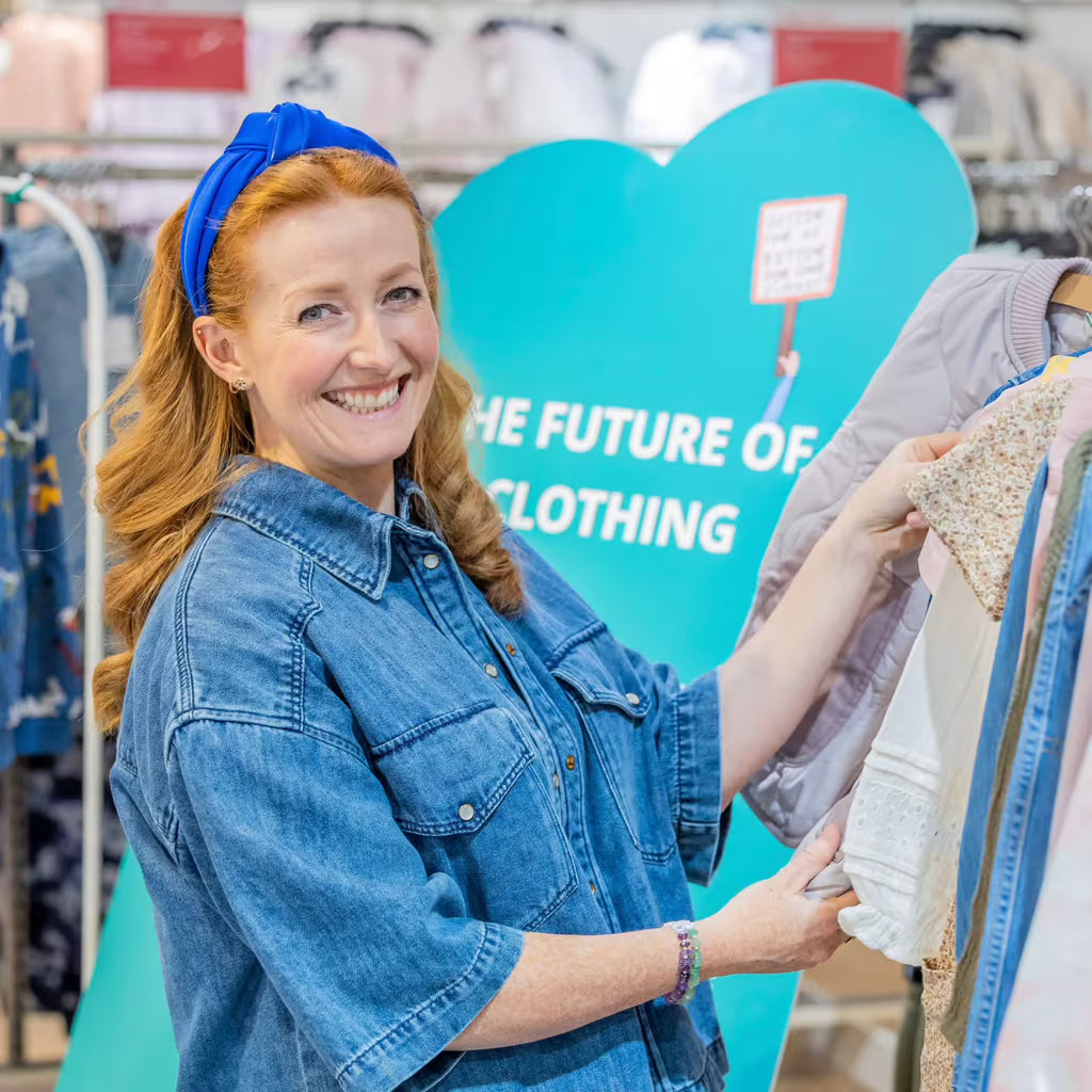 Woman in a denim shirt holding clothing in front of a heart-shaped sign with text in a store.