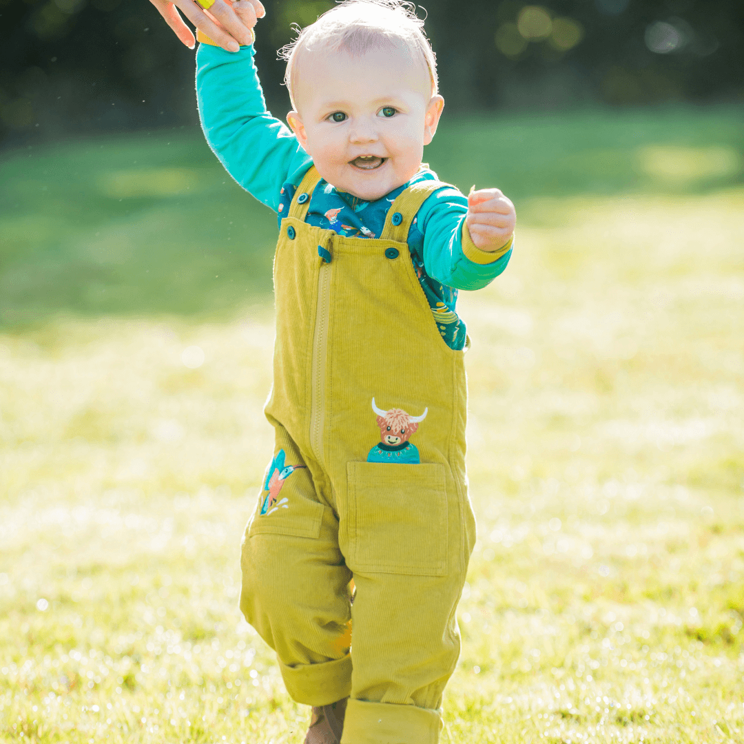 Baby in green outfit standing on grass with adult hand holding