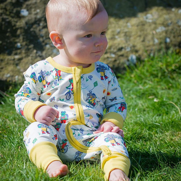Baby sitting in the grass wearing a zip-up unisex sleepsuit with frogs, hippos and bees walking and cycling together