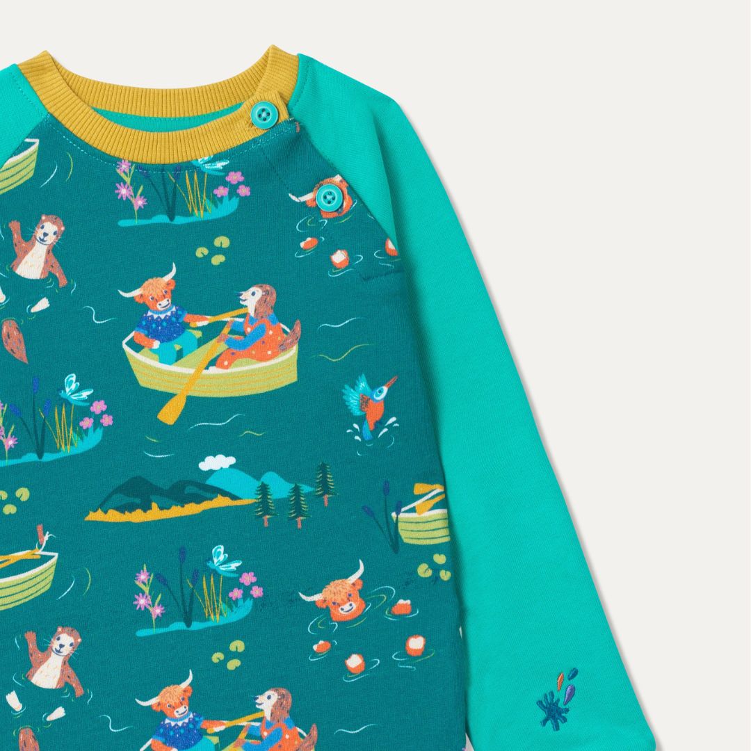 Close up of teal sweatshirt with a fun repeat print of a highland cow, otter and king fisher wild swimming and rowing. The sweatshirt has teal sleeves, a mustard yellow trim and two turquoise buttons on the shoulder. 