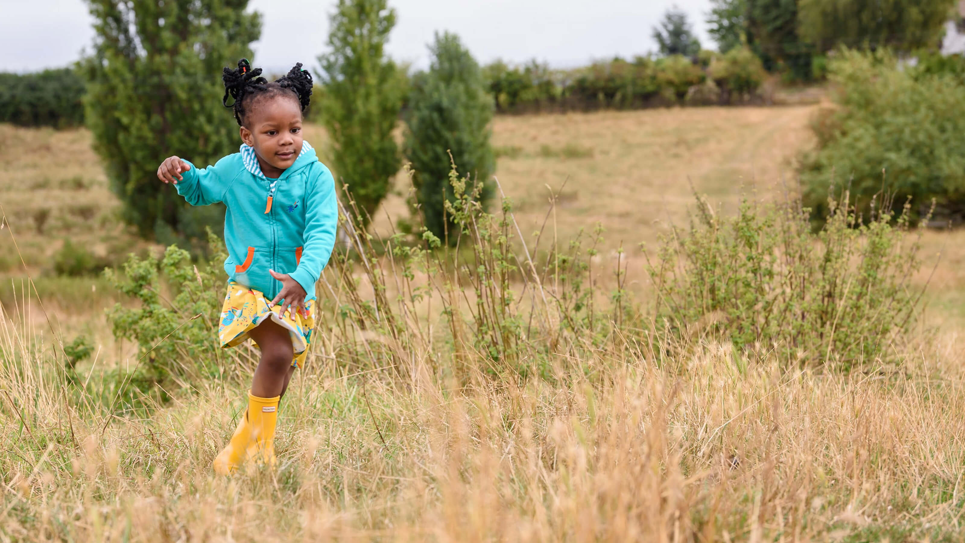 Child in a blue jacket and yellow rain boots running through tall grass in a field.