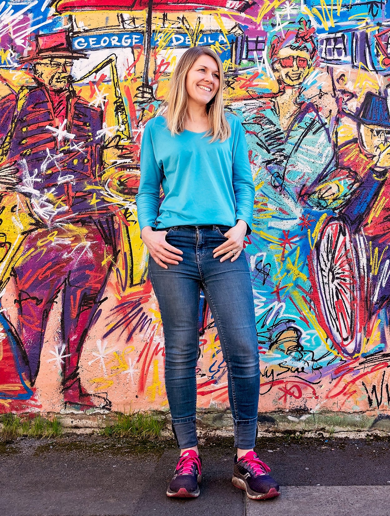 Woman in a blue shirt and jeans standing in front of a colorful graffiti wall.