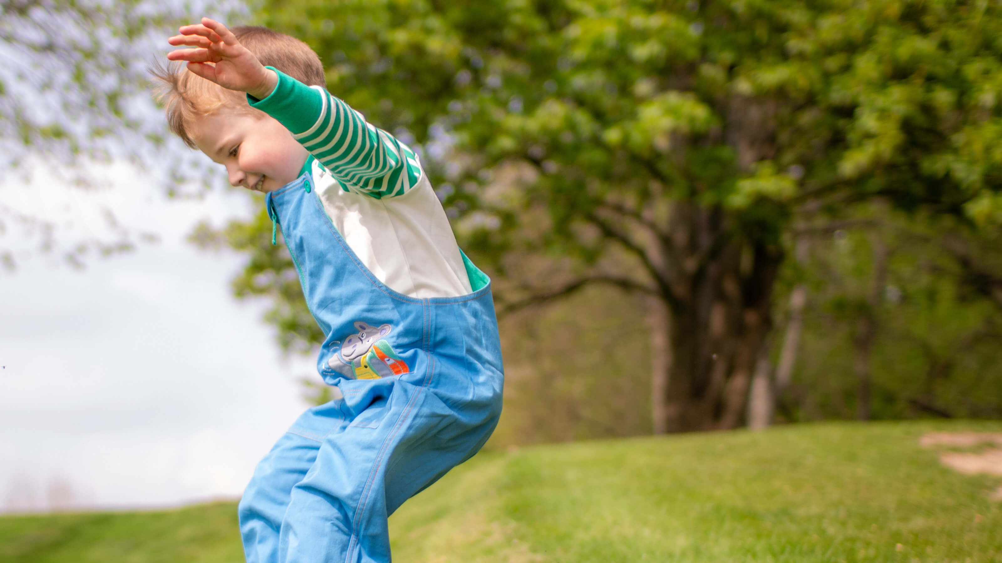 Child in blue overalls and green sweater playing outdoors with trees and grass in the background