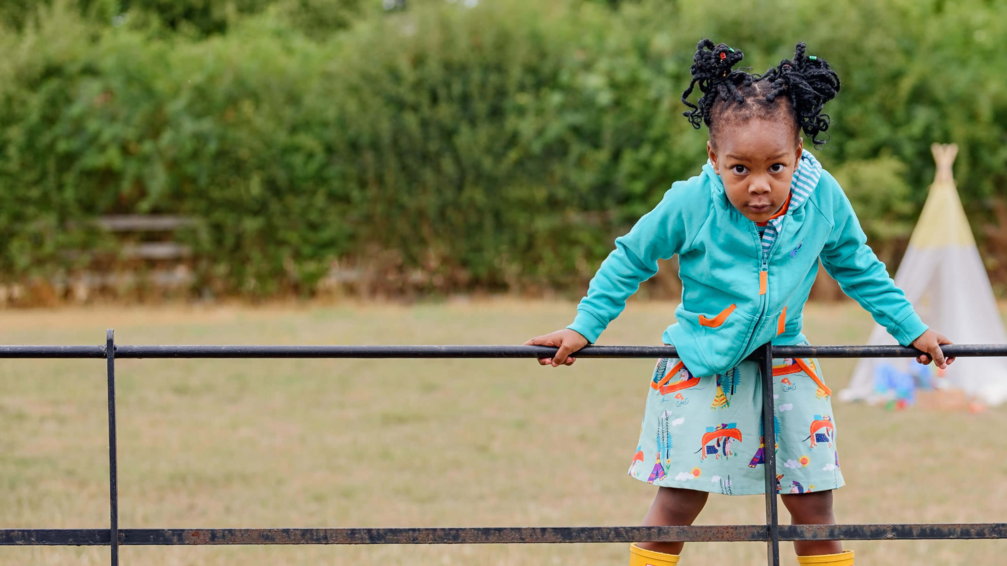 Child in a teal jacket and colorful skirt standing on a metal fence in an outdoor setting.