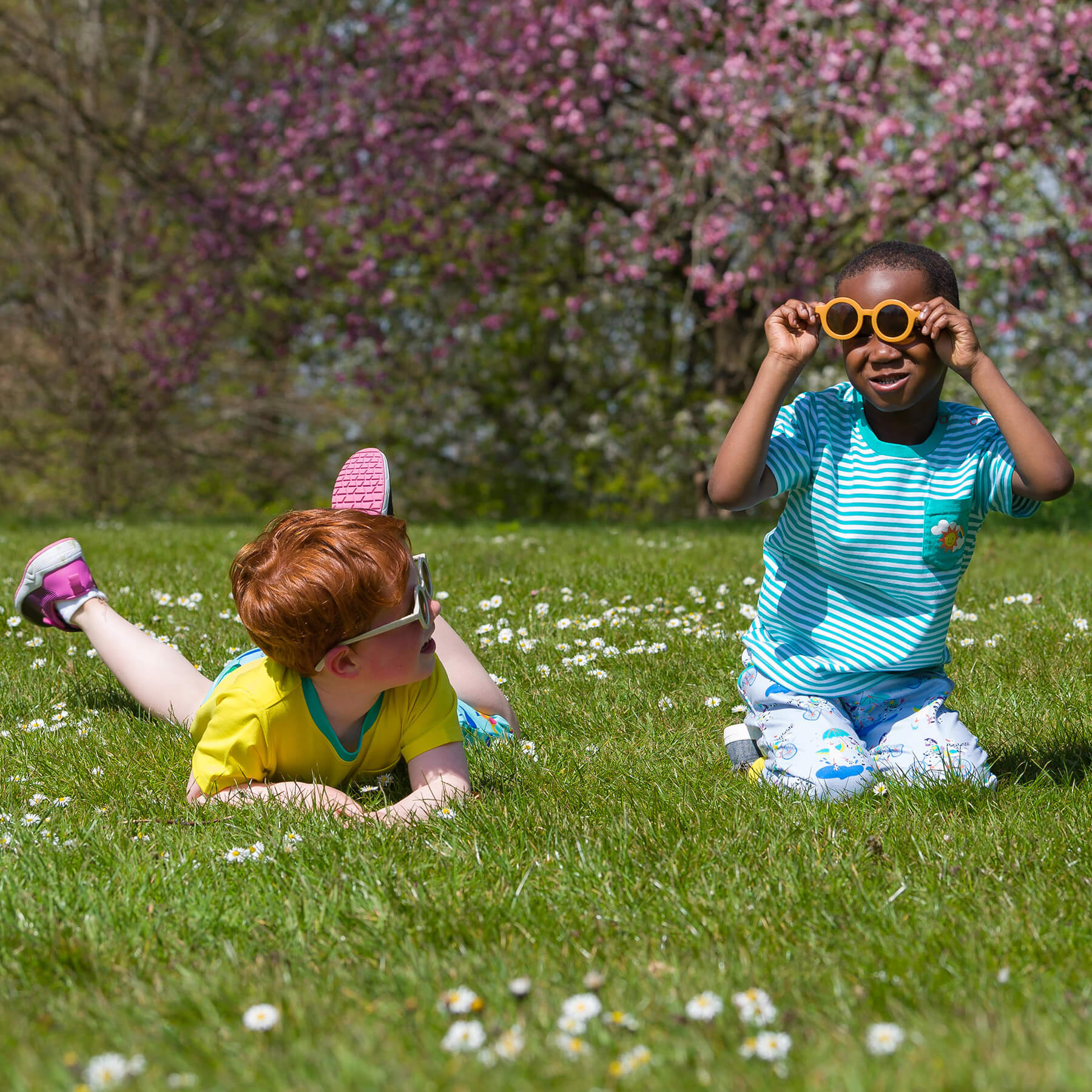Two children playing in a grassy area with flowers and a flowering tree in the background.