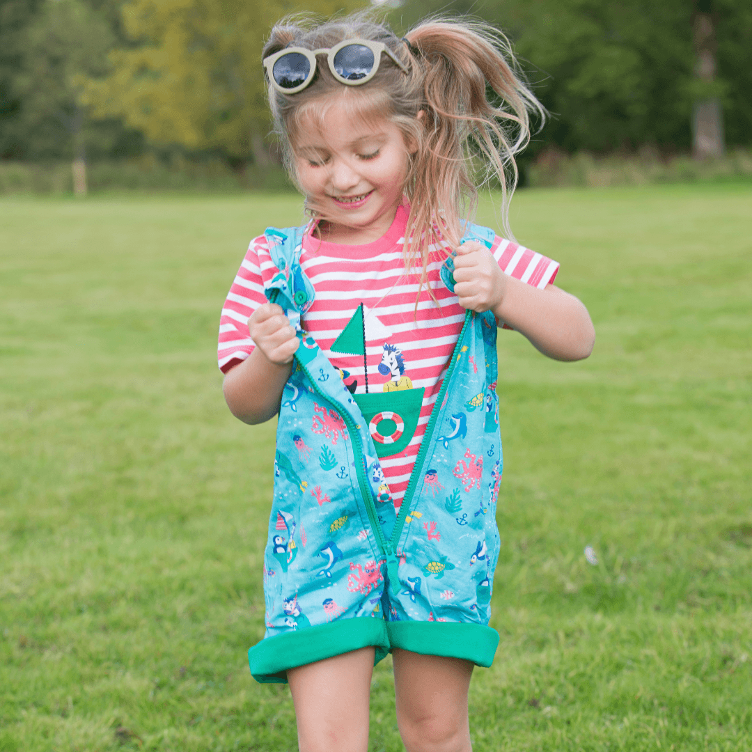 Smiling girl in her Ducky Zebra coral pink t-shirt and ocean-themed dungarees with a green zip. The girl is outside, and has opened her zip to show the t-shirt
