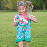 Smiling girl in her Ducky Zebra coral pink t-shirt and ocean-themed dungarees with a green zip. The girl is outside, and has opened her zip to show the t-shirt