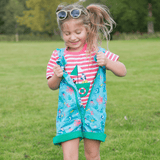 Smiling girl in her Ducky Zebra coral pink t-shirt and ocean-themed dungarees with a green zip. The girl is outside, and has opened her zip to show the t-shirt