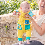 Image of a baby being held up outside. The baby is wearing yellow dungarees with turquoise pockets. One pocket has an applique of a sun and cloud. The other has an applique of a hot air balloon popping out of the pocket. 