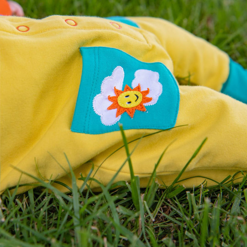 Close up image of a baby wearing yellow dungarees. The photo focuses on its turquoise pocket. The pocket has an applique of a sun and clouds.