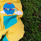 Close up image of a baby wearing a yellow sleeveless romper. The photo focuses on its turquoise pocket. The pocket has a hot air balloon and rainbow applique pocking out of it.  