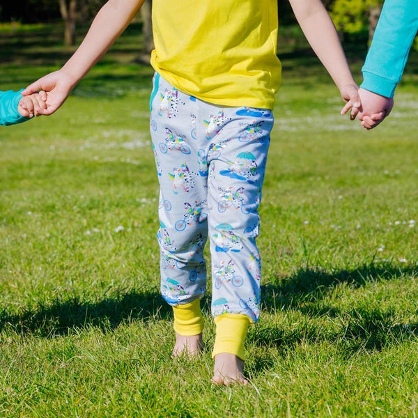 Image of a child wearing a pair of brightly coloured joggers with a light grey background and repeat print pattern of a duck and zebra splashing in puddles, riding a bike and running a three legged race. The child is holding hands with two other children and is walking with bare feet through the grass