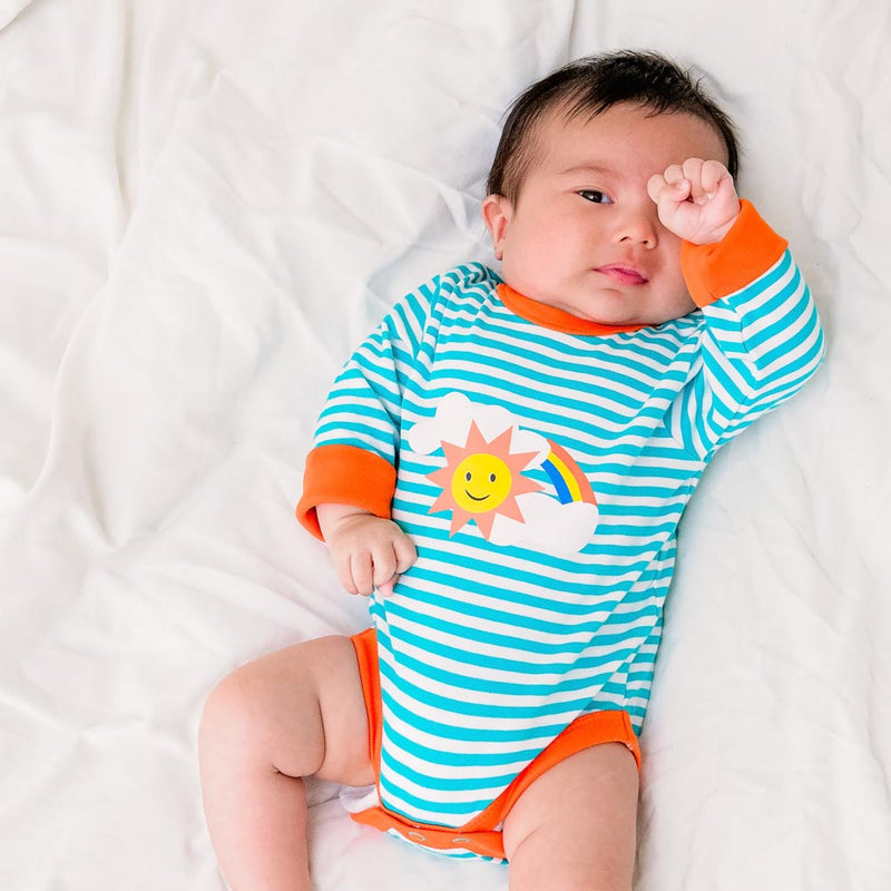 Image of a tired baby rubbing their eyes, wearing a long sleeve turquoise stripe bodysuit with flame orange trim and roll-up arm cuffs. The bodysuit has a sun, rainbow and cloud print on the chest