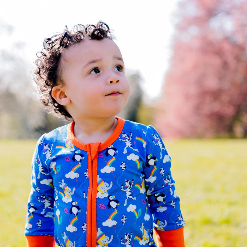 Image of an inquisitive toddler, wearing a long sleeve zip-up unisex baby romper with a bright blue background and colourful repeat print pattern of an elephant, zebra and puffin flying a kite. The image shows flame orange cuffs, trim and a two way zip