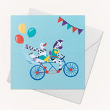 Image of a square greeting card with a picture of a duck and zebra cycling on a tandem bicycle, with balloons blowing behind them. An envelope is tucked behind the card.