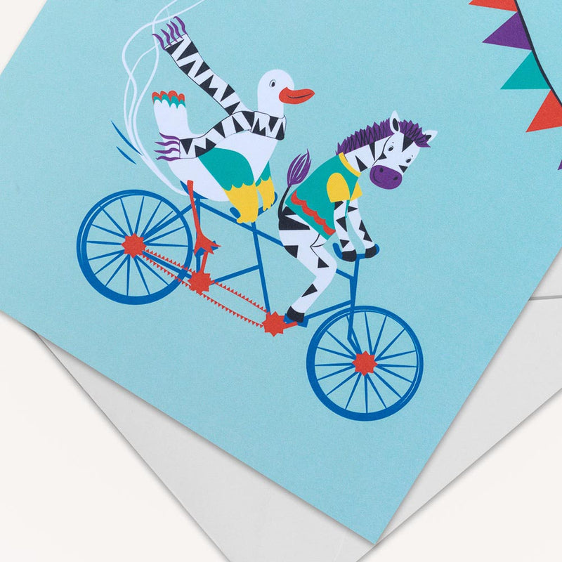 Image of a square greeting card with a picture of a duck and zebra cycling on a tandem bicycle, with balloons blowing behind them