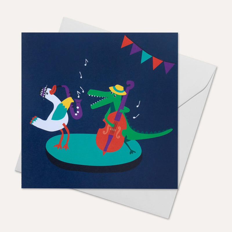 Square, navy blue greeting card with a duck and crocodile playing the saxophone and double bass together. An envelope is sitting behind the card.