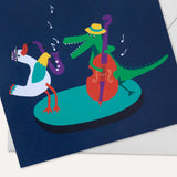 A close up image of a square, navy blue greeting card with a duck and crocodile playing the saxophone and double bass together. An envelope is sitting behind the card.