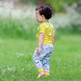 Image of a toddler walking away from the camera, wearing a pair of brightly coloured joggers with a light grey background and repeat print pattern of a duck and zebra splashing in puddles, riding a bike and running a three legged race. The child has bare feet and is walking through the grass.