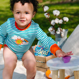 Image of a smiling baby playing outside, wearing a long sleeve turquoise stripe bodysuit with flame orange trim and roll-up arm cuffs. The bodysuit has two turquoise poppers on the neck and a sun, rainbow and cloud print on the chest