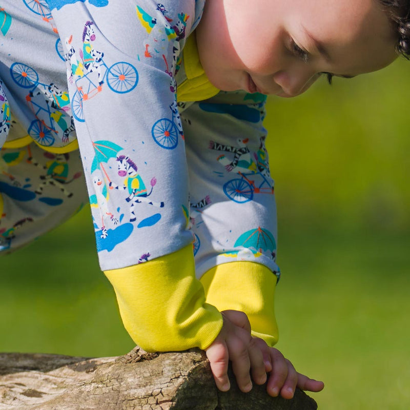 Image of a confident baby balancing on a log, wearing a long sleeve zip-up unisex baby romper with a light grey background and colourful repeat print pattern of a duck and zebra splashing in a puddle, riding a bicycle and running a three legged race. The images shows bright yellow cuffs, trim and a two way zip