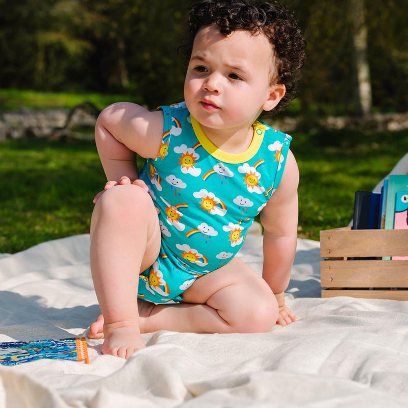 Image of a baby wearing a unisex sleeveless baby bodysuit with a turquoise background and repeat print pattern of rainbows, sun and clouds. The image shows the front of the bodysuit with a yellow neck trim and two turquoise poppers on the shoulder