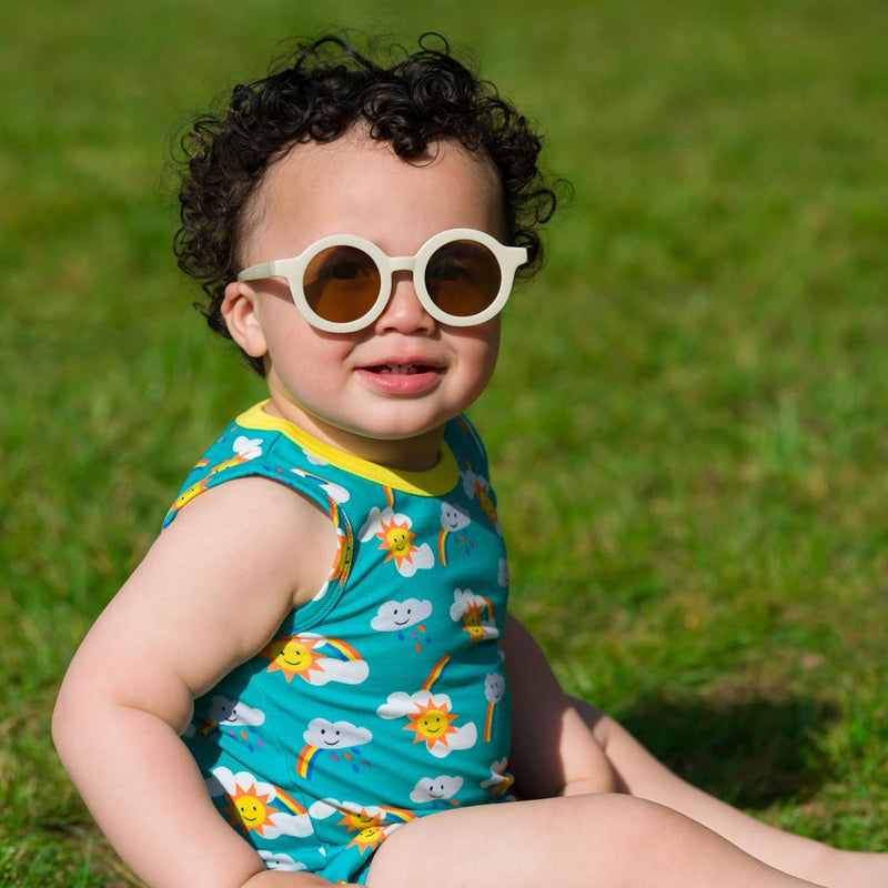 Image of a smiling baby, wearing white sunglasses and a sleeveless Ducky Zebra bodysuit. The unisex bodysuit has a turquoise background and repeat print pattern of rainbows, sun and clouds.