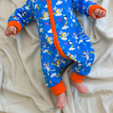Image of a baby wearing a long sleeve zip-up unisex baby romper with a bright blue background and colourful repeat print pattern of an elephant, zebra and puffin flying a kite. The image shows flame orange cuffs, trim and a two way zip