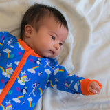 Image of a baby wearing a long sleeve zip-up unisex baby romper with a bright blue background and colourful repeat print pattern of an elephant, zebra and puffin flying a kite. The image shows flame orange cuffs, trim and a two way zip