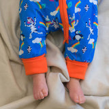 Image of a baby wearing a long sleeve zip-up unisex baby romper with a bright blue background and colourful repeat print pattern of an elephant, zebra and puffin flying a kite. The image focuses on the bottom half of the sleepsuit, with the flame orange cuffs, trim and a two way zip