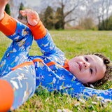 Image of a happy toddler rolling in the grass, wearing a long sleeve zip-up unisex baby romper with a bright blue background and colourful repeat print pattern of an elephant, zebra and puffin flying a kite. The image shows flame orange cuffs, trim and a two way zip