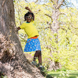 Image of a smiling child leaning against a tree wearing a Ducky Zebra yellow t-shirt and colourful, kite flying repeat print skort