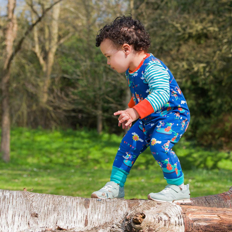 Image of a toddler balancing on a log wearing a Ducky Zebra sleeveless romper and long sleeve bodysuit. The dungarees have a repeat print pattern of a duck, crocodile, cheetah and fox playing in a festival.