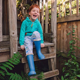 Image of a smiling boy sitting in a tree house wearing Ducky Zebra trousers and a hoodie with wellies
