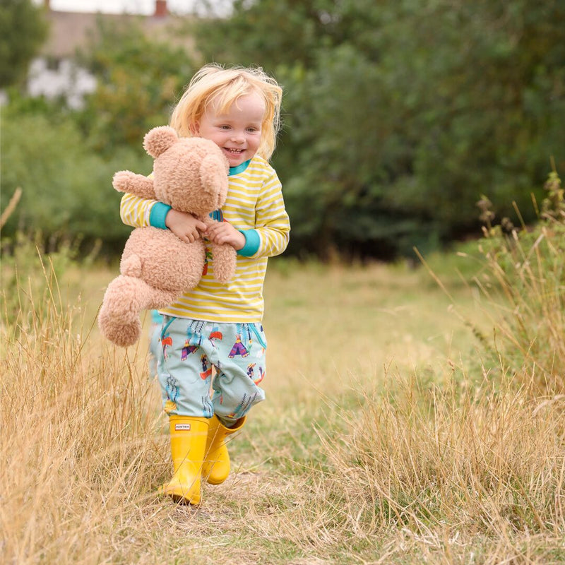 Image of a girl running through the grass, cuddling a teddy and wearing a pair of Ducky Zebra green trousers and a yellow and white stripe Ducky Zebra long sleeve top. She's wearing a pair of yellow wellies.