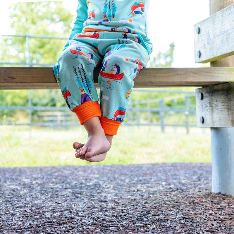 Image of a child's legs dangling underneath a table. The child is wearing colourful Ducky Zebra green joggers with a canoeing dog and hedgehog print and orange cuff.