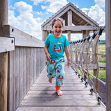 Image of a happy girl running across a wooden bridge in a playground wearing a pair of Ducky Zebra green joggers with a canoeing dog and hedgehog print