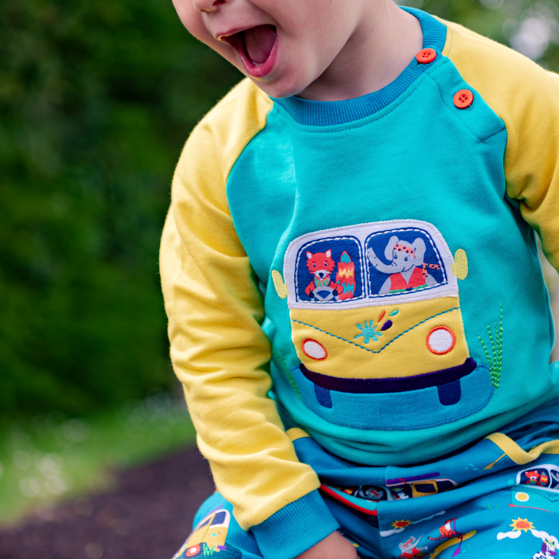 Image of a child wearing a colourful Ducky Zebra jumper. The jumper has a picture of a fox and elephant driving in a yellow campervan together. The jumper has yellow sleeves, teal cuffs and a turquoise main body. 